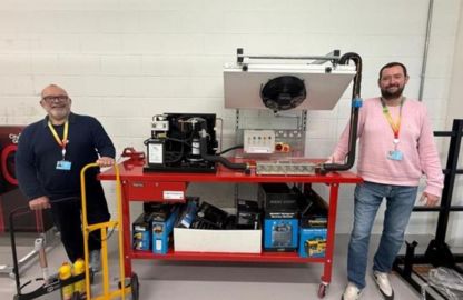 Two men stand smiling beside a red workbench filled with tools and equipment. A cooling system is on the table. A friendly and collaborative atmosphere.
