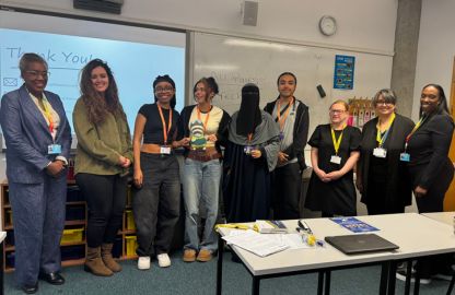 A diverse group of nine people stand smiling in a classroom. Behind them are a whiteboard and projector screen. The setting is professional and friendly.