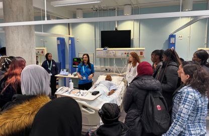 Students observe a lecturer demonstrating nursing techniques at a hospital bed surrounded by a group of attentive learners.